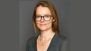 Professional headshot of Kirsten Kastberg, G&rsquo;26, wearing glasses and a dark top, smiling against a gray background.