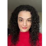 Headshot of Olivia Barrows, smiling at the camera. She has shoulder-length dark curly hair and is wearing a bright red turtleneck sweater against a neutral gray background.