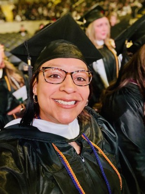 Elizabeth Broady smiling in a graduation cap and gown, wearing glasses and honor cords, seated among other graduates during a commencement ceremony.