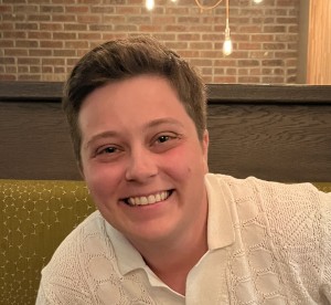 &ldquo;Grayson Connery smiling while seated, wearing a light-colored collared shirt, with a brick wall and hanging lights in the background