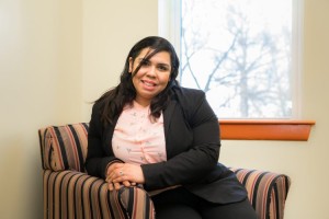 Nayroby Rosa smiling while seated in a striped armchair, wearing a black blazer over a light pink blouse, with a window and trees visible in the background.