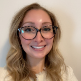 Headshot of Shana Dulac, smiling at the camera. She has long blonde wavy hair, wears large dark-framed glasses and a cream-colored top.
